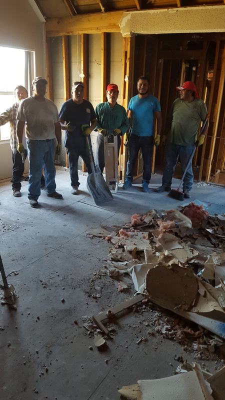 Six workers with shovels standing in a room stripped to the studs, with torn drywall and insulation on the floor
