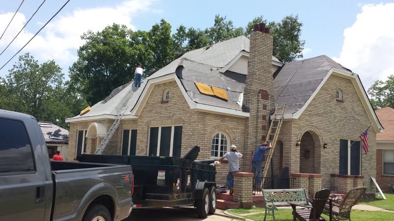 Crew removing shingles from the roof of a brick cottage with a chimney, a ladder against the wall and a dump trailer parked in front